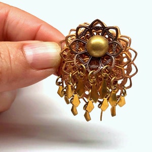 May include: A vintage gold-toned brooch with a floral filigree design. The brooch features a central sphere and dangling diamond-shaped charms. The intricate metalwork and warm tones create a classic, elegant aesthetic.
