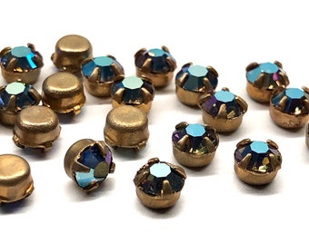 May include: A group of iridescent blue rhinestones with gold settings. The rhinestones are scattered on a white surface.