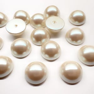 May include: A collection of off-white, half-round pearl cabochons with a smooth, lustrous finish. Each piece features a domed top and a flat base, with some showing a small central hole on the underside, arranged in a scattered pattern.