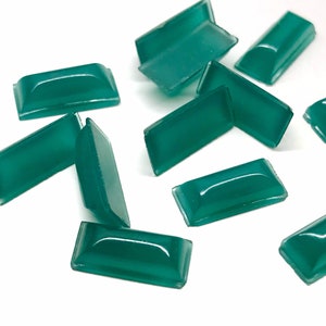 May include: A group of teal green rectangular shaped glass cabochons. The cabochons are scattered on a white surface.