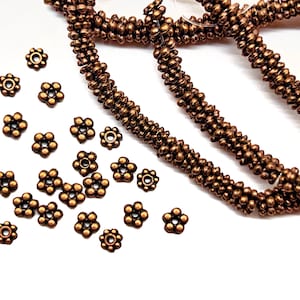 May include: A collection of small, copper-colored metal flower-shaped beads with a central hole, scattered alongside a long, continuous strand of identical, closely spaced, textured copper-colored cylindrical beads on a white surface.