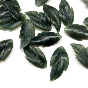 May include: Dark green jade leaf carvings. Each leaf is intricately carved with detailed veining, creating a realistic appearance. The leaves are scattered on a white surface, showcasing their natural color and texture. Ideal for jewelry making or decorative purposes.