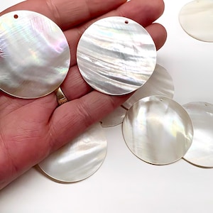 May include: A collection of circular mother of pearl shell pendants, each featuring a smooth, iridescent surface with natural pearlescent white tones and a single small hole at the top for hanging, showcasing a polished finish.