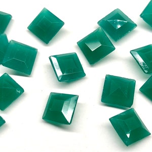 May include: Emerald green square-cut gemstones scattered on a white surface. Each gem has a faceted design, catching the light. The gems are approximately 0.5 inches in size, suitable for jewelry making or crafts.