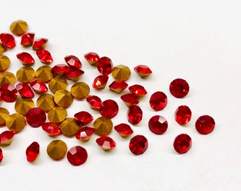 May include: A scattering of red and gold rhinestones on a white surface. The red rhinestones are faceted and round, while the gold ones have a faceted, geometric design. Ideal for craft projects.