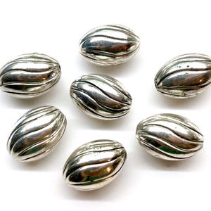 May include: Seven oval-shaped beads with a metallic silver finish. Each bead features a series of raised, diagonal ridges that wrap around the surface, creating a textured, ribbed appearance. The beads are arranged in a loose cluster.