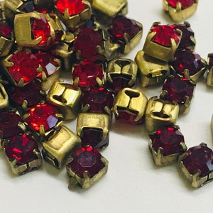 May include: A pile of red glass rhinestones with gold metal settings. The rhinestones are square shaped and have a faceted design.