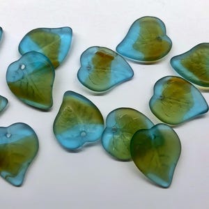 May include: A collection of leaf-shaped glass beads. Each bead has a translucent design with a gradient of colors, including blue, green, and amber. The beads have a hole for stringing and are scattered on a white surface.