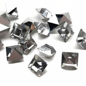 May include: A scattering of clear, faceted, square-shaped rhinestones on a white background.