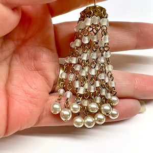 May include: A decorative tassel ornament featuring multiple brass-toned metal chains suspended from a circular brass cap. Each chain is adorned with small, translucent white glass beads and finished with a larger, opaque white pearl-like bead.