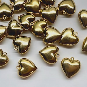 May include: A collection of small, shiny gold-toned heart charms, each featuring a smooth, rounded surface and a single metal loop at the top for attachment. These metallic heart-shaped pendants are scattered across the frame in various orientations.