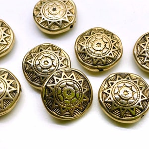 May include: Close-up of several gold-colored, round beads with a sunburst design. Each bead has a central floral pattern surrounded by a starburst design. The beads are slightly irregular in shape and have a metallic finish.