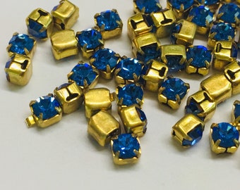May include: A scattering of blue rhinestone embellishments with gold settings. The rhinestones are square shaped and have a faceted design. The settings are square shaped and have a small loop on the top.