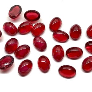 May include: A collection of oval-shaped, deep red glass beads. The beads are scattered across a white surface, each displaying a glossy, translucent quality. The color varies from a bright ruby to a darker, almost burgundy hue.