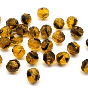 May include: A collection of faceted amber-colored beads with dark brown swirls. The beads are scattered on a white surface, showcasing their geometric shapes and translucent quality. Ideal for jewelry making.