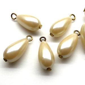 May include: Six off-white pearlescent teardrop-shaped beads with gold-toned metal loops. The beads are approximately 1 inch in length.