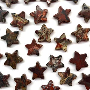 May include: A collection of star-shaped gemstones in various shades of red, brown, and tan. The stones have a natural, speckled appearance and are arranged against a white background. The stars are approximately 1 inch in size.