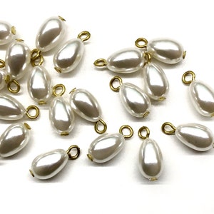 May include: A collection of white pearl teardrop shaped beads with gold-toned metal loops. The beads are scattered on a white surface.