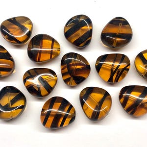 May include: A collection of amber-colored, tumbled glass beads with black stripe designs. The beads are oval and irregular in shape, with a glossy finish. The beads are arranged on a white surface.
