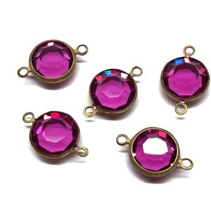 May include: Five round, faceted magenta-colored crystal connectors with brass-colored metal settings and loops. The crystals have a brilliant shine, and the connectors are arranged on a white background.