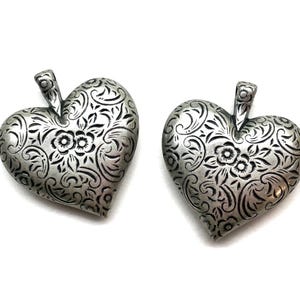 May include: Two silver-toned heart-shaped pendants with intricate floral and swirl patterns. Each pendant has a small loop at the top for hanging. The pendants are displayed on a white background.