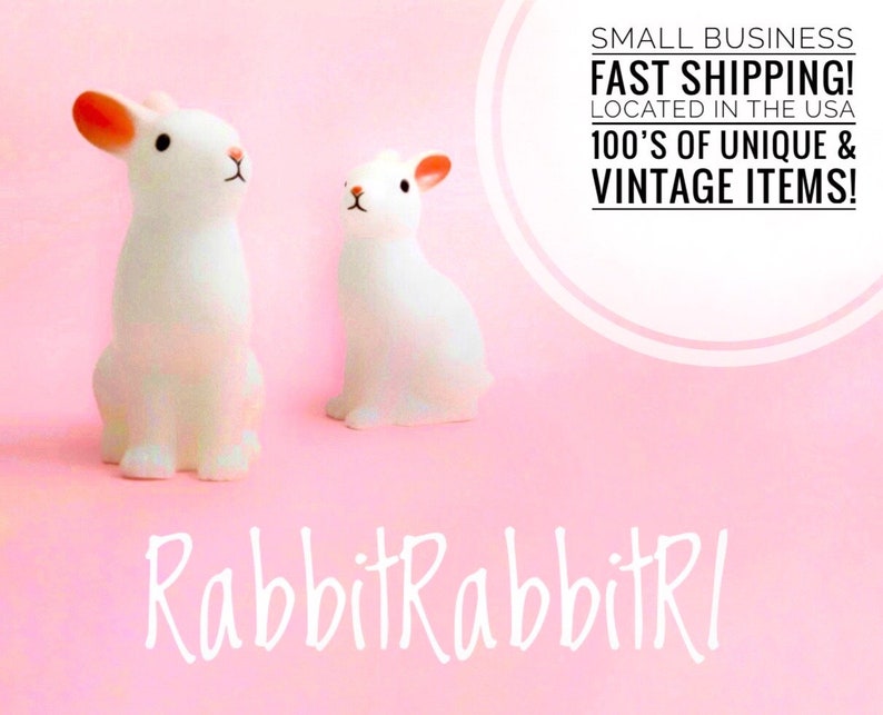 May include: Two white ceramic rabbit figurines on a pink background. The larger rabbit has pink ears and is sitting upright. The smaller rabbit is sitting with its head tilted to the side. The text "RabbitRabbitRl" is written in white cursive letters below the rabbits.