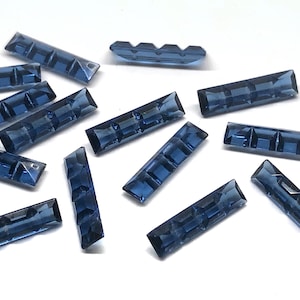 May include: A collection of dark blue, faceted, rectangular glass rhinestones. The rhinestones are scattered on a white surface.