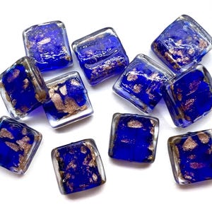 May include: A collection of ten square glass beads featuring a deep cobalt blue base color with embedded metallic copper-colored flakes throughout each piece. The beads have a smooth, glossy finish and a uniform square shape with rounded edges.