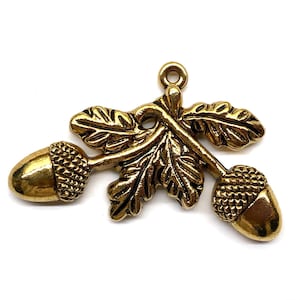 May include: A gold-colored metal charm featuring two acorns and leaves. The acorns have textured caps and are connected to the leaves by stems. The charm has a loop at the top for attaching to jewelry or other crafts.