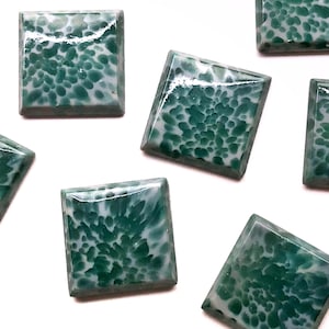 May include: Close-up of several square-shaped, green and white speckled gemstones. The gemstones have a glossy finish and are arranged on a white surface. The green and white pattern resembles a natural stone.