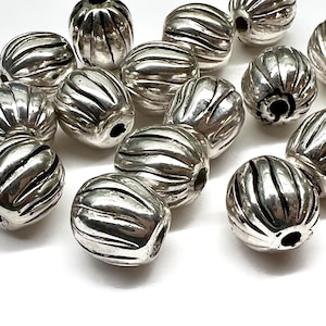 May include: A collection of round, silver-colored beads featuring a vertical ribbed texture. Each bead has a central hole for stringing, with a polished metallic finish and dark recessed lines between the raised ridges across the surface.