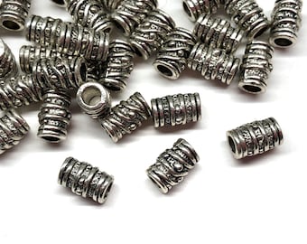 May include: Silver-colored metal beads with a cylindrical shape and intricate etched designs. The beads have a weathered, antique appearance and are scattered on a white surface. These beads are suitable for jewelry making and crafts.