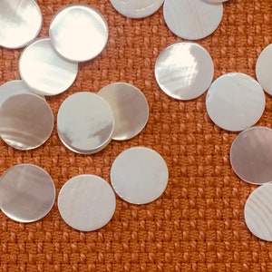 May include: A collection of iridescent, round, mother-of-pearl discs scattered on an orange woven surface. The discs vary in size and reflect light, creating a shimmering effect. These decorative elements could be used for crafts or embellishments.