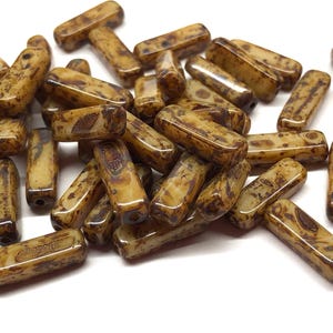 May include: A collection of rectangular beads with a mottled pattern in shades of brown and tan. The beads have a glossy finish and are scattered across a white surface. Each bead is approximately 0.5 inches long.