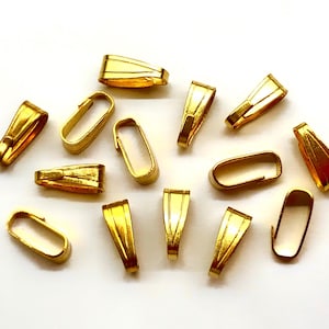 May include: A collection of gold-colored jewelry bails in various shapes, including rectangular and trapezoidal designs. The bails are arranged on a white surface, showcasing their shiny, metallic finish. These findings are used for jewelry making.