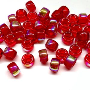 May include: A collection of translucent red beads with a glossy finish. Some beads have a slight iridescent sheen, reflecting gold and purple hues. The beads are various shapes, including rounded and cylindrical, suitable for jewelry making.