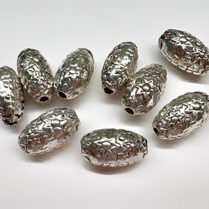 May include: A collection of nine oval-shaped silver-toned beads, each featuring a repeating embossed star and floral pattern across the entire surface. The beads have a metallic finish and a central hole running through the length of each piece.