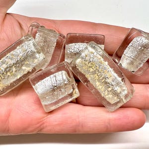 May include: A collection of five clear glass cabochons, each featuring a rectangular or square shape with an embedded layer of metallic silver foil and small, textured beige inclusions visible through the transparent glass surface.
