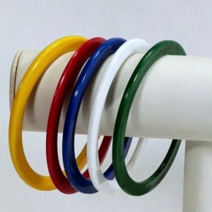 May include: Five colorful plastic bangles in yellow, red, blue, white, and green. The bangles are stacked on a white display stand.
