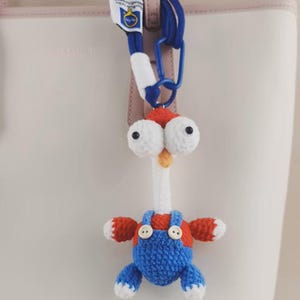 May include: A crocheted keychain featuring a character with a long white neck, large white eyes with black pupils, and a small orange beak. The character wears blue overalls with two white buttons over a red shirt and has white-tipped limbs.