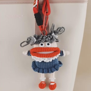 May include: A handmade crochet doll keychain with a whimsical design. The doll features a large orange mouth, blue dress, and red shoes. The keychain is attached to a red and black carabiner clip, perfect for bags or keys.