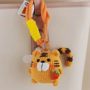Puede incluir: Un llavero de tigre tejido a ganchillo con forma redonda, hecho de hilo naranja, rayas negras, hocico blanco y ojos negros. El tigre sostiene una pequeña fresa roja y tiene una cola a rayas. Está sujeto a un clip de plástico naranja con un detalle de hilo amarillo.