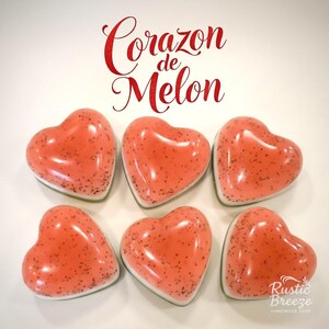 May include: Six heart-shaped soaps with a speckled, coral-colored appearance. The soaps have a glossy finish and a white base. The image includes the text "Corazon de Melon" in red script and the "Rustic Breeze Handmade Soap" logo.