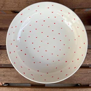 May include: A white ceramic serving bowl with a glossy finish. The bowl is decorated with a pattern of small, evenly spaced red hearts. The bowl is round and has a slightly curved rim. The bowl is sitting on a wooden surface.