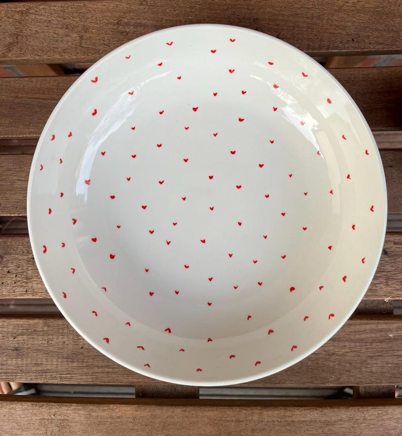 May include: A white ceramic serving bowl with a glossy finish. The bowl is decorated with a pattern of small, evenly spaced red hearts. The bowl is round and has a slightly curved rim. The bowl is sitting on a wooden surface.