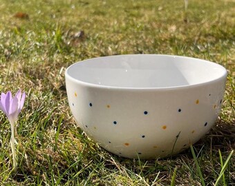 以下が含まれることがあります： 青と黄色の水玉模様の白いセラミックボウルが草の上に置かれています。ボウルは丸く、シンプルなデザインです。背景には草と小さな紫色の花が写っています。