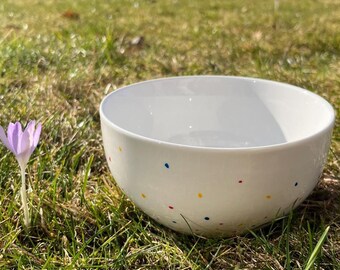 以下が含まれることがあります： 赤、黄、青のカラフルな水玉模様が施された白いセラミックボウル。ボウルは緑の草の上に置かれ、前景には紫色のクロッカスが咲いています。ボウルは丸く、滑らかで光沢のある仕上がりです。