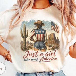 Puede incluir: Camiseta de color marfil con un gráfico de una mujer con sombrero de vaquero, ropa con temática de la bandera estadounidense y un paisaje desértico. El texto "Just a girl who loves America" está en escritura roja. También se ve un cactus y la bandera estadounidense.