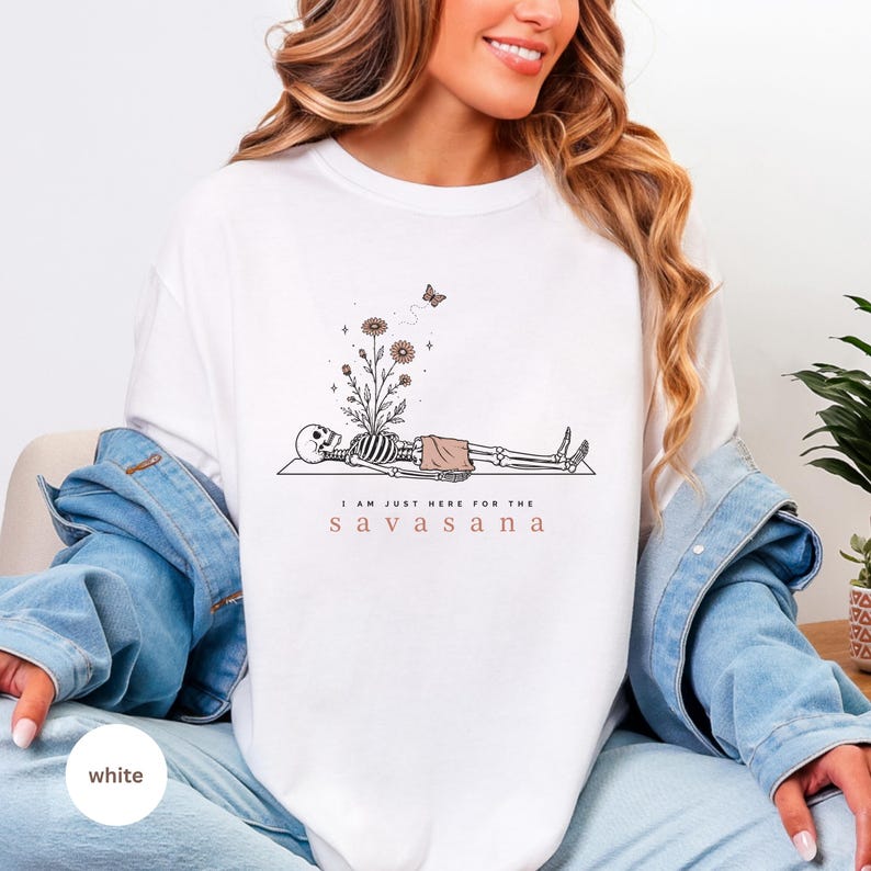 Puede incluir: Una sudadera blanca de cuello redondo con un gr&aacute;fico de l&iacute;neas negras de un esqueleto acostado en una esterilla de yoga, con flores creciendo de su pecho y una mariposa encima. El texto dice I AM JUST HERE FOR THE SAVASANA en negro y rosa.