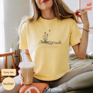 Puede incluir: Una camiseta de manga corta de color amarillo p&aacute;lido con un gr&aacute;fico de l&iacute;neas negras de un esqueleto en pose de yoga, con flores creciendo de su pecho y una peque&ntilde;a mariposa encima. El texto bajo el gr&aacute;fico dice I AM JUST HERE FOR THE SAVASANA.